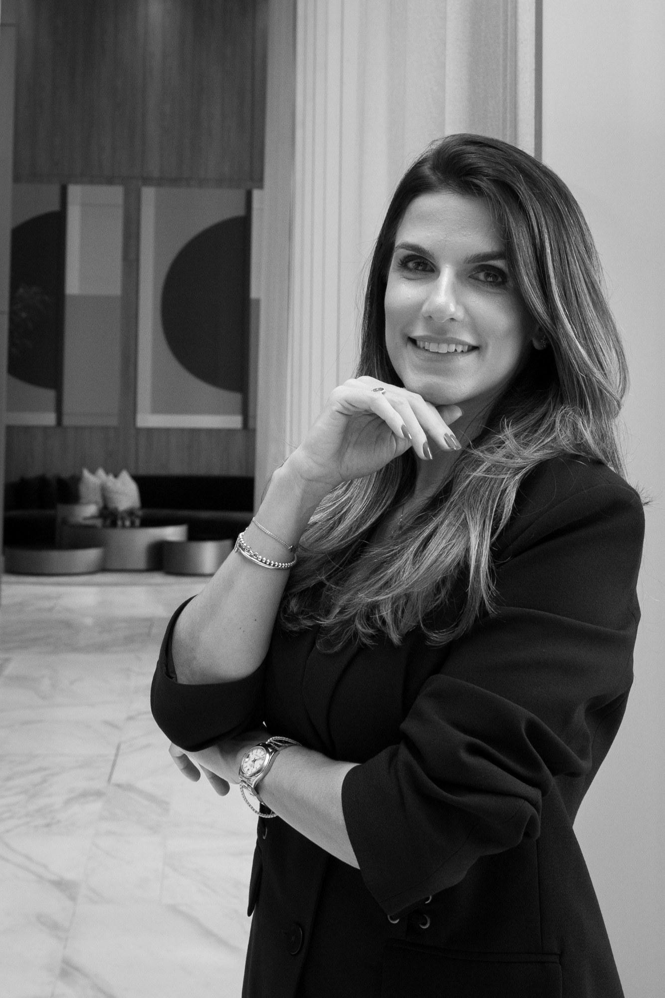 Retrato em preto e branco de Carolina Monnerat, sorrindo, com a mão apoiada no queixo em um lobby.