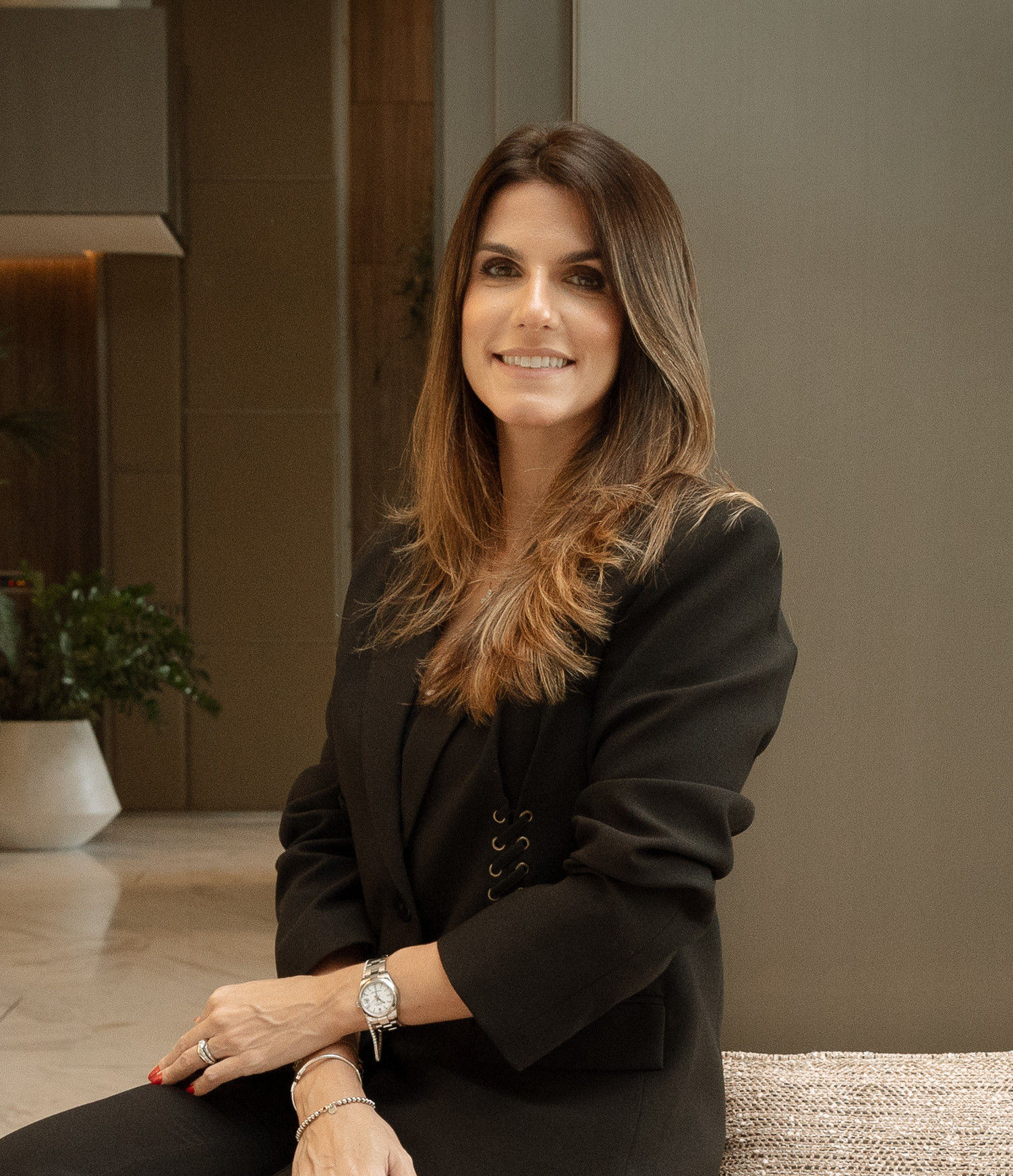 Carolina Monnerat sorrindo, sentada no braço de um sofá, com blazer preto, em um lobby com piso de mármore e plantas.